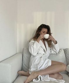 a woman is sitting on a couch drinking from a cup while wrapped in a white robe