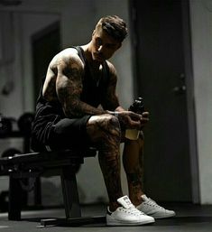 a man with tattoos sitting on a bench holding a bottle and looking at his cell phone