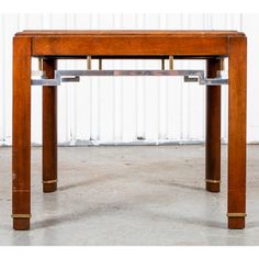 a wooden table with two metal legs and a square top on concrete flooring in front of a white wall