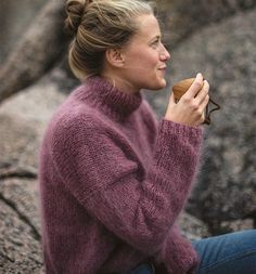 a woman is sitting on the rocks and holding a cup in her hand while wearing a purple sweater