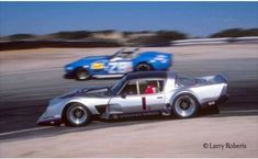 two race cars racing on a track during the day, one is silver and the other is blue