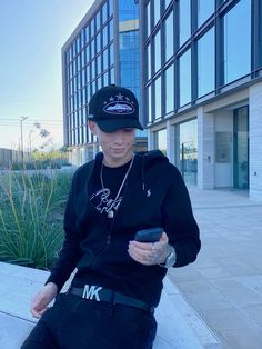 a young man sitting on the ground looking at his cell phone while wearing a hat