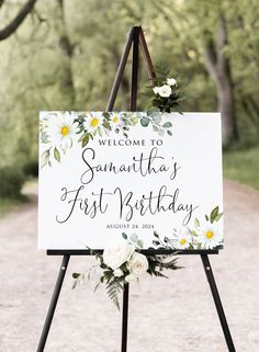 a welcome sign with flowers and greenery is displayed on an easel in the middle of a dirt road