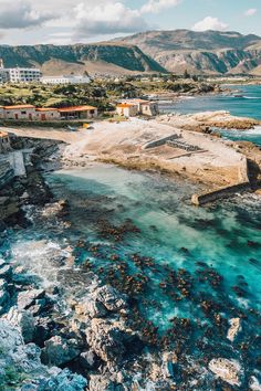 an aerial view of the ocean and rocky coastline with buildings in the distance, surrounded by mountains