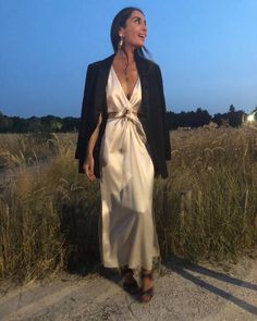 a woman in a white dress and black blazer is standing on the sand near tall grass