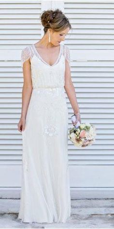 a woman standing in front of a white wall wearing a dress and holding a bouquet