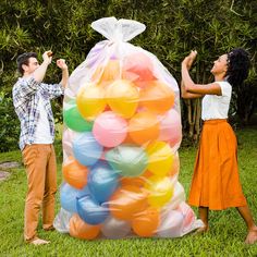 a man and woman standing next to a large bag filled with balloons in the grass