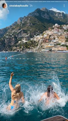 two people are swimming in the ocean near a town and mountains with houses on it