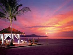 a gazebo sitting on top of a sandy beach under a palm tree at sunset