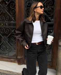 a woman in black jacket and jeans holding a coffee cup standing next to a building