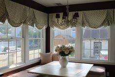 a vase with flowers on top of a table in front of two windows and a bench