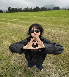 a person standing in the grass making a heart sign with their hands and wearing sunglasses