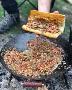 a person holding a sandwich over an open fire in the ground with other food on it