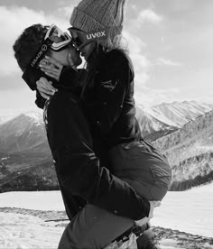 a man and woman kissing on top of a snow covered ski slope with mountains in the background