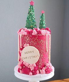 a pink and green decorated christmas cake on a white pedestal with two trees in the background