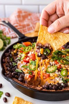 a person dipping a tortilla chip into a skillet filled with black beans, cheese and veggies