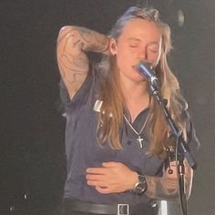 a woman with long hair standing in front of a microphone and holding her hands behind her head