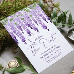 purple flowers and greenery are on the ground next to a save the date card