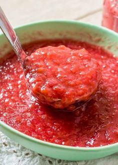 a spoon full of red sauce in a green bowl with another container behind it on the table