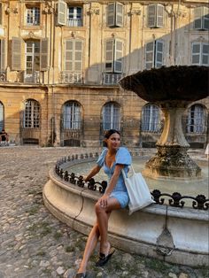 a woman sitting on the edge of a fountain in front of an old building with lots of windows