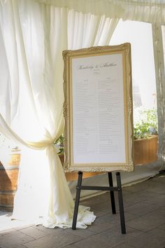 a white and gold framed wedding program on a easel in front of a drape