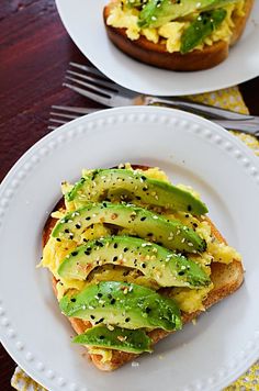 two white plates topped with sandwiches covered in avocado and other toppings on top of a wooden table