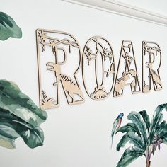 the word roar written in wooden letters on a white wall with tropical leaves and plants