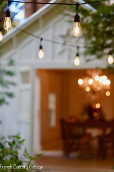 some lights are hanging over a table and chairs in front of a house with an open door