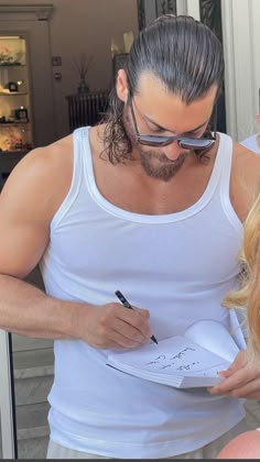 a man in white tank top writing on paper next to a blonde haired woman wearing sunglasses