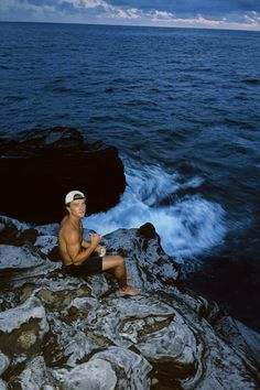 a man sitting on top of a rocky cliff next to the ocean holding a cup