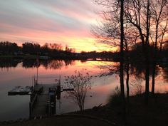 the sun is setting over a lake with a dock and trees in front of it