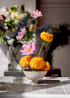 three flowers in a white bowl on a table with vases behind it and another flower arrangement in the background