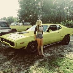 a woman standing next to a yellow car in a field with other cars behind her