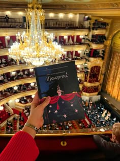 a person holding up a book in front of an auditorium filled with people and chandeliers