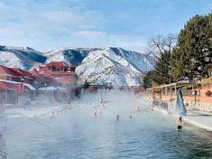 many people are in the water near buildings and snow covered mountains, with steam rising from them