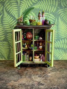 a green cabinet filled with lots of items on top of a table next to a fern leaf wallpaper