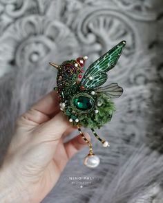 a hand holding a tiny green fairy brooch with pearls and crystals on it's wings