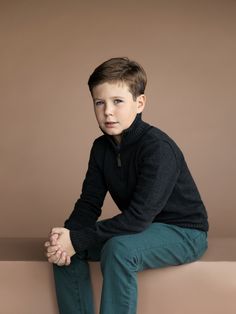 a young boy sitting on top of a bench wearing green pants and a black sweater