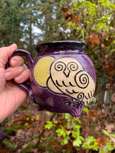 a hand holding a purple and yellow owl mug in front of some trees with green leaves