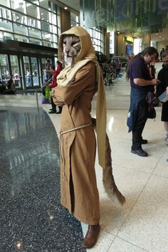 a man in a trench coat and mask standing next to an airport lobby area with people walking around