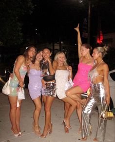 four beautiful young women standing next to each other on a sidewalk at night with their arms in the air