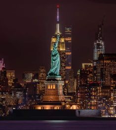 the statue of liberty is lit up at night in new york city, with skyscrapers behind it