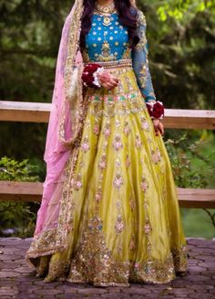 a woman in a yellow and blue lehenga is standing on a bridge with trees behind her