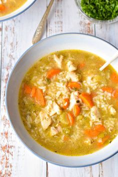 two bowls of chicken and rice soup on a white wooden table with carrots, parsley