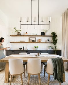 a dining room table with white chairs and a woman in the background walking past it