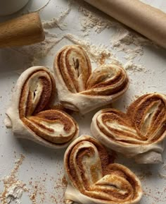 four cinnamon roll shaped like hearts sitting on top of a white surface next to a rolling pin