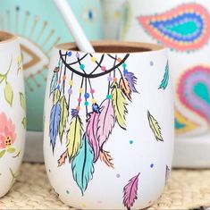 two cups with feathers painted on them and one has a spoon in the cup next to it