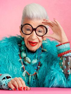 an older woman wearing glasses and a blue fur coat with beads around her neck, sitting at a table in front of a pink background