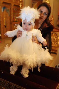 a woman holding a baby wearing a white dress and feathered headdress in front of a mirror