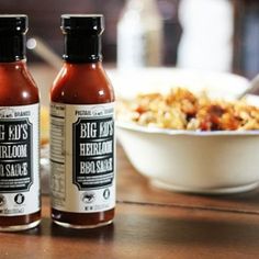 two bottles of bbq sauce sit on a table next to a bowl of food